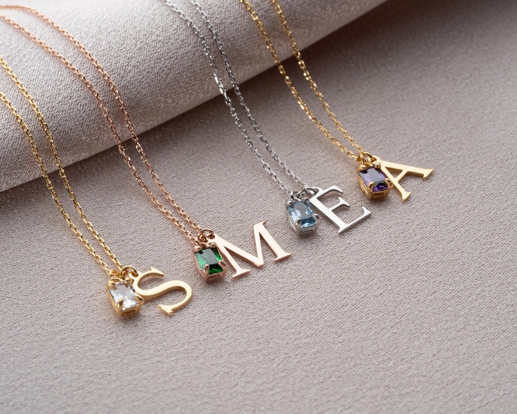 A high-quality 5:4 product hero shot of multiple gold, silver, and rose gold initial necklaces featuring colorful gemstone accents.