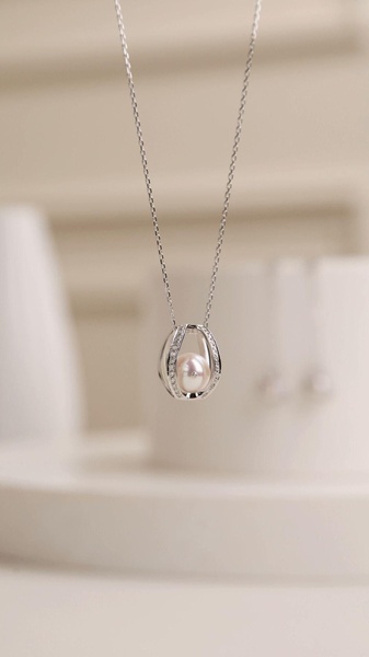 A high-end, clean-background capture of a delicate pearl pendant necklace suspended in a decorative, cage-like metal frame.