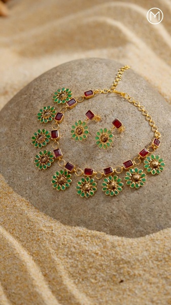This creative showcases a gold necklace with floral-shaped green and red gems, elegantly placed on a natural stone surrounded by beach sand.