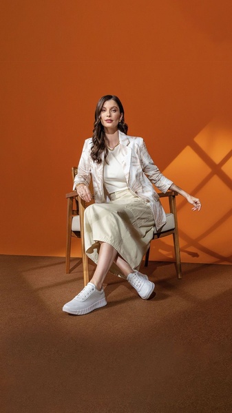 A high-quality, studio-lit photograph featuring a woman in smart-casual attire wearing clean white sneakers while seated in a modern chair.