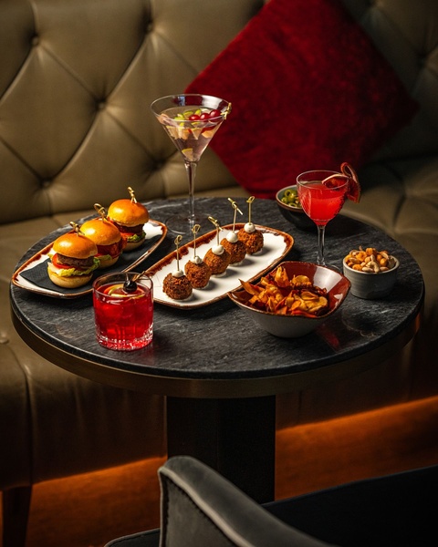 A sophisticated lifestyle shot featuring gourmet sliders and appetizers paired with craft cocktails on a marble table in a dim, intimate setting.