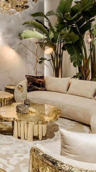 A high-end interior design shot featuring a curved cream sofa, a metallic gold organic-shaped coffee table, and large indoor tropical plants.
