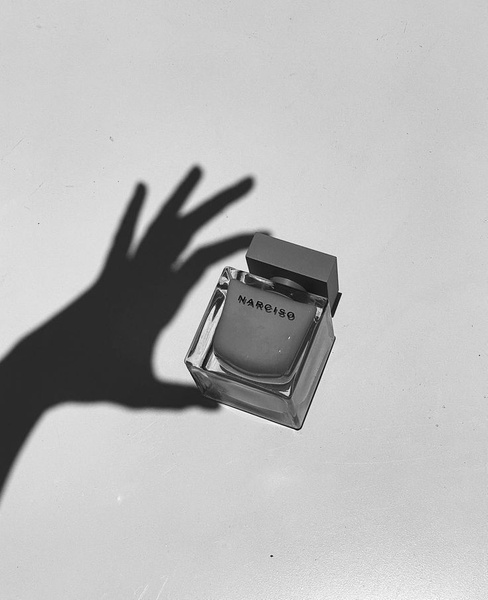 A high-contrast, black and white product shot of a square perfume bottle being reached for by a silhouetted hand, creating an air of mystery and desire.