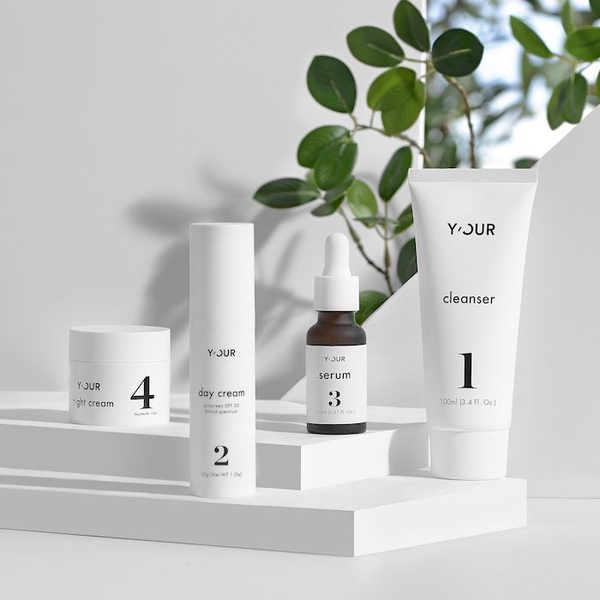 A clean, high-key studio photograph featuring a four-piece skincare set arranged on tiered pedestals with natural foliage in the background.