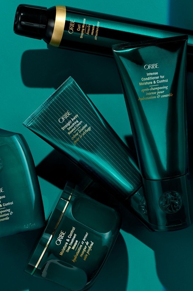 A high-end editorial display featuring a curated collection of hair care bottles and tubes arranged in a monochromatic deep teal environment.