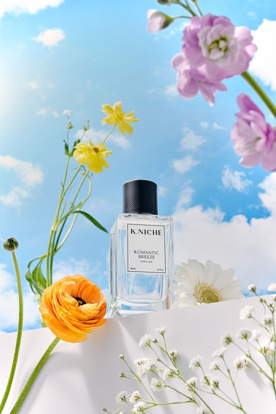 A high-end, bright, and airy product hero shot of a K.Niche perfume bottle centered among colorful spring flowers against a clear, sunny blue sky.