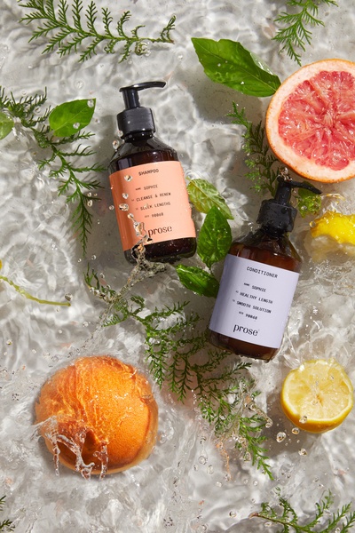 A high-end, clean beauty advertisement featuring shampoo and conditioner bottles submerged in water with fresh citrus and botanical elements.