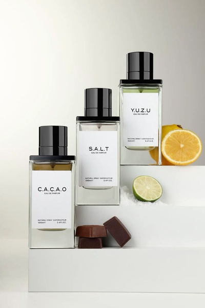 A clean, high-end product photography shot featuring three perfume bottles labeled C.A.C.A.O, S.A.L.T, and Y.U.Z.U, arranged on tiers with raw ingredient accents.