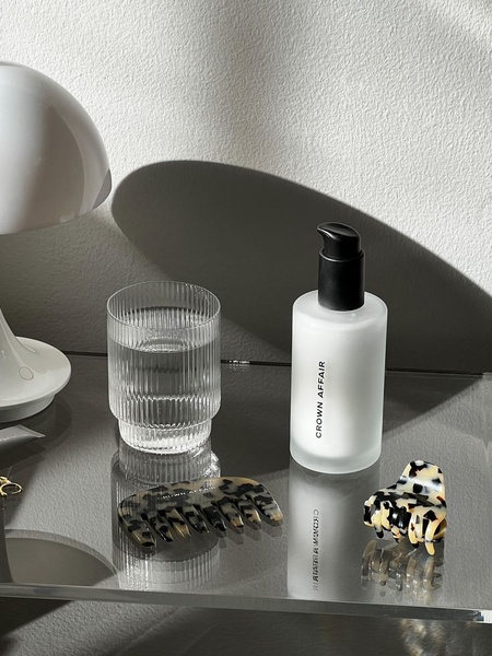 A clean, high-end product hero shot featuring a bottle of hair serum next to hair accessories and a glass of water on a reflective surface.