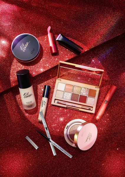 A top-down, high-contrast flat lay of various makeup products including cushions, palettes, and brushes set against a deep red glitter background.