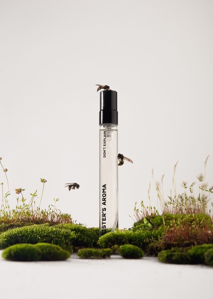 A minimalist product shot featuring a perfume bottle placed among real moss and small wildflowers with bees resting on the bottle and flying nearby.
