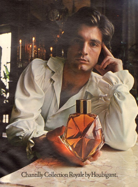 A high-end perfume advertisement featuring a man in a period-style shirt holding a faceted glass fragrance bottle in a candlelit, moody setting.