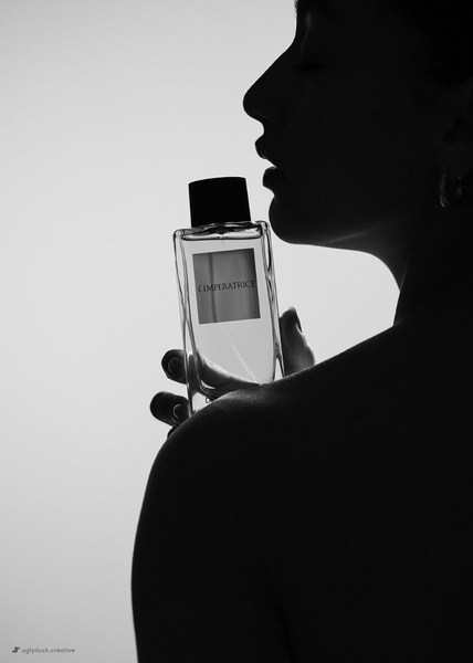 A high-contrast, black and white silhouette shot highlighting a perfume bottle against a soft, backlit profile of a woman.
