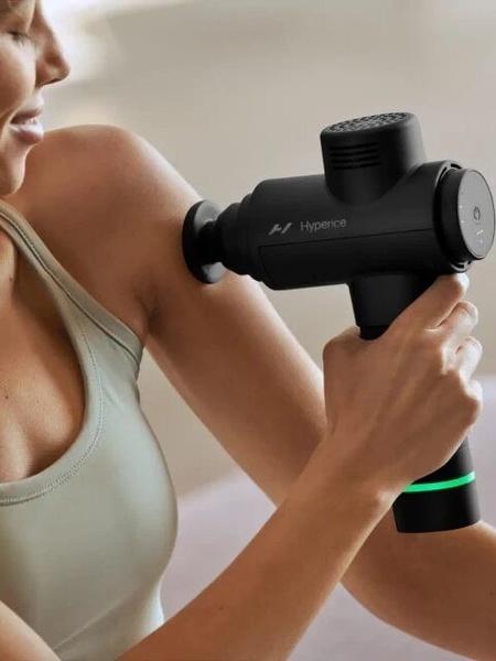 A close-up shot of an individual using a massage gun on their shoulder, highlighting the device's ergonomic design and clean interface.