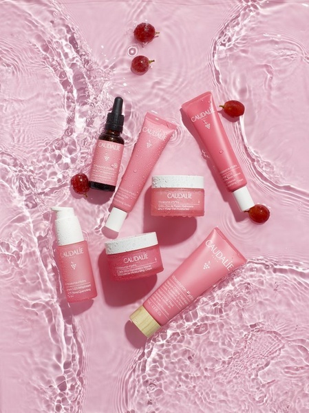 A top-down, clean-beauty product photography shot showcasing a range of skincare bottles and creams submerged in ripples of water with fresh grapes.