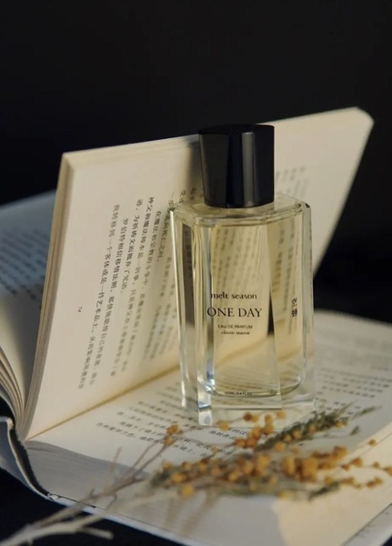 A close-up, editorial-style product shot featuring a perfume bottle placed atop an open book, accompanied by dried flowers against a dark background.