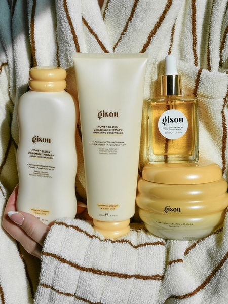A high-end, minimalist product hero shot featuring a full collection of hair care bottles and jars held against a textured backdrop.