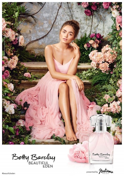 A perfume product hero shot featuring a model in a light pink dress surrounded by a lush floral garden backdrop.