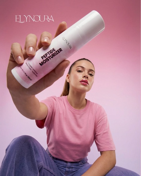 A close-up shot of a model holding a branded peptide moisturizer bottle toward the camera against a solid pink backdrop.
