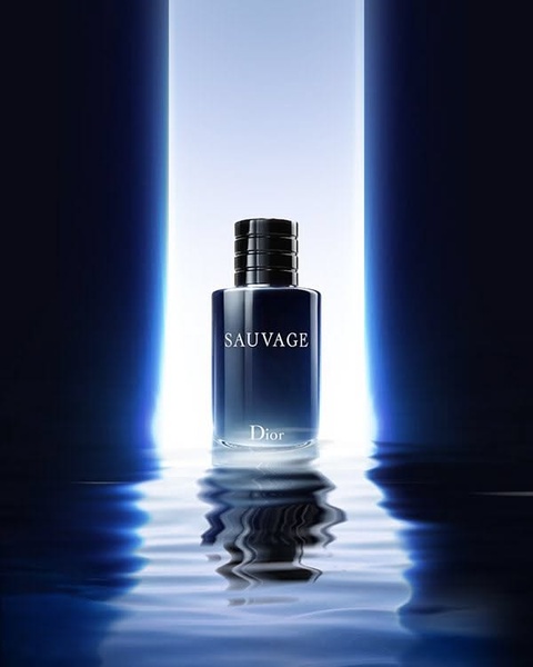 A single bottle of perfume is centered against a bright, vertical light beam, with a clear and crisp reflection appearing on the water surface below.