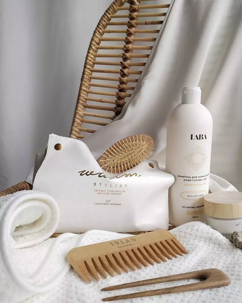 A high-end, clean product photography composition featuring shampoo, a wooden comb, and a hair pin against a textured rattan backdrop.