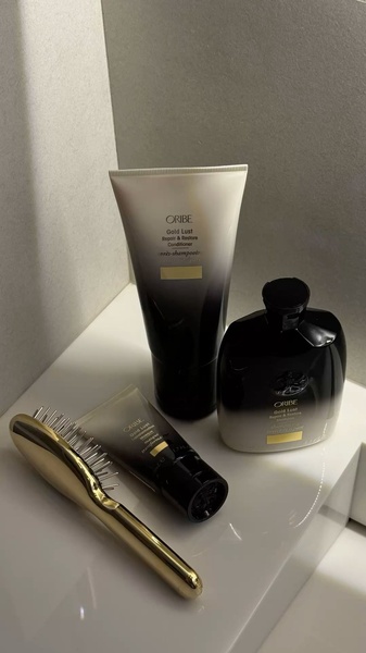 A high-end, studio-lit shot of Oribe Gold Lust hair care products arranged on a clean, modern surface with a gold brush.