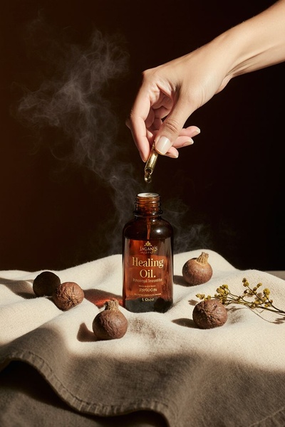 A close-up shot showing a hand using a glass dropper to apply healing oil onto a textured cloth, surrounded by natural botanical elements.