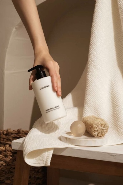 A high-end, clean-toned product photograph of a body wash pump bottle placed next to a loofah and soap stone on a textured towel.