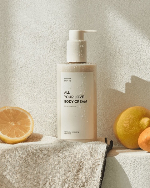 A high-quality, minimalist product shot of a body cream bottle positioned next to natural lemons on a textured towel, conveying a fresh and organic brand feel.