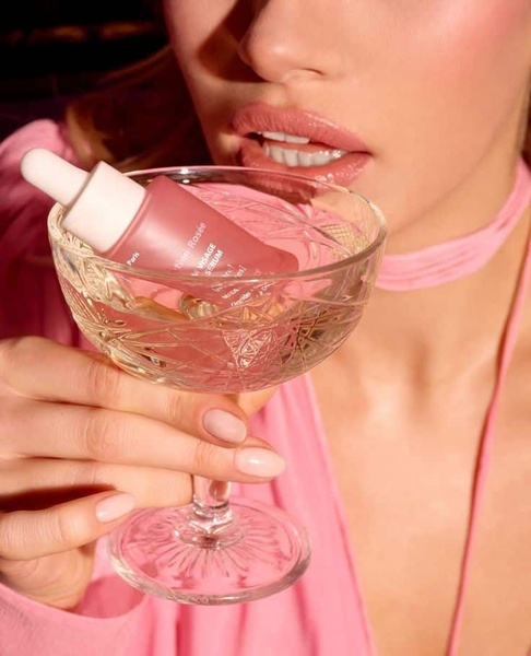 A close-up aesthetic shot of a pink serum bottle nestled inside an elegant, faceted cocktail glass held by a model with subtle, matching makeup.