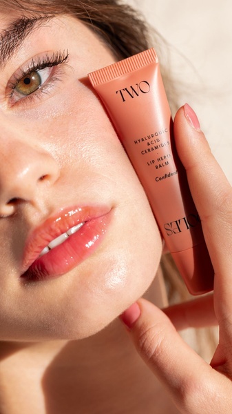 A high-quality 9:16 vertical close-up showing a model holding a lip balm tube next to her glossy, hydrated lips.