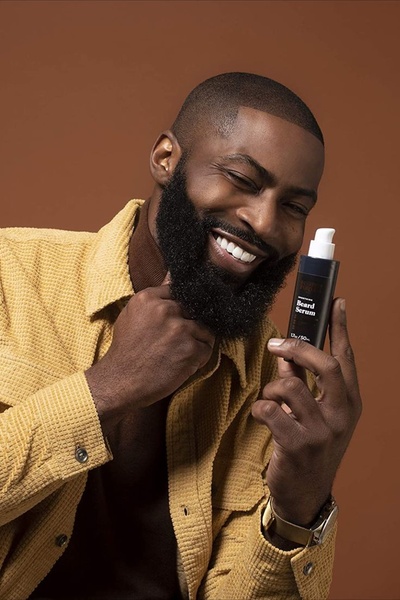 A high-quality portrait of a smiling man with a thick, well-groomed beard, showcasing a Beard Serum bottle to highlight product results and personal confidence.