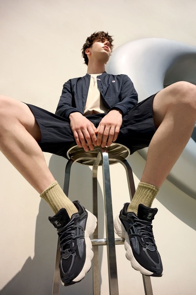 A high-fashion, low-angle studio shot focusing on the footwear, featuring a model sitting on a stool to emphasize the design and texture of the running shoe.