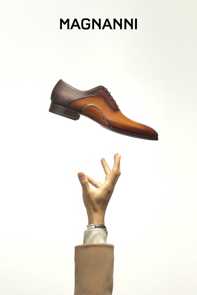 A high-end leather dress shoe appears to be floating above a reaching hand, highlighting product craftsmanship through minimal, clean studio lighting.