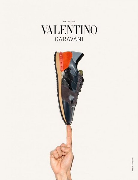 A high-fashion running shoe is balanced perfectly on the tip of a human finger against a clean, off-white background.
