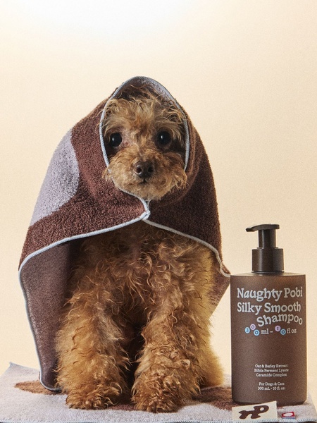 A high-quality studio shot of a dog wrapped in a towel next to a bottle of Naughty Pobi shampoo, emphasizing a clean and premium aesthetic.