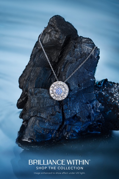 A high-end diamond necklace is draped over a jagged, dark charcoal rock, set against a calming blue, water-like background to emphasize jewelry sparkle.
