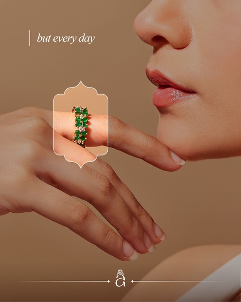 A close-up photograph of a model wearing a delicate emerald and diamond ring, featuring a soft-focus background and elegant framing.