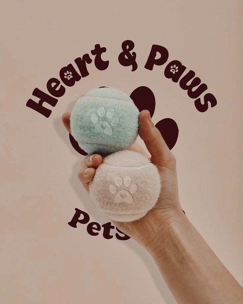 A close-up shot of two textured tennis balls for pets, held against a soft-toned background featuring a paw print graphic.