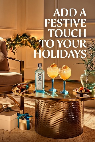 A static ad featuring a bottle of Bombay Sapphire gin next to two garnished cocktails on a gold table, set against a warm, holiday-themed living room background.