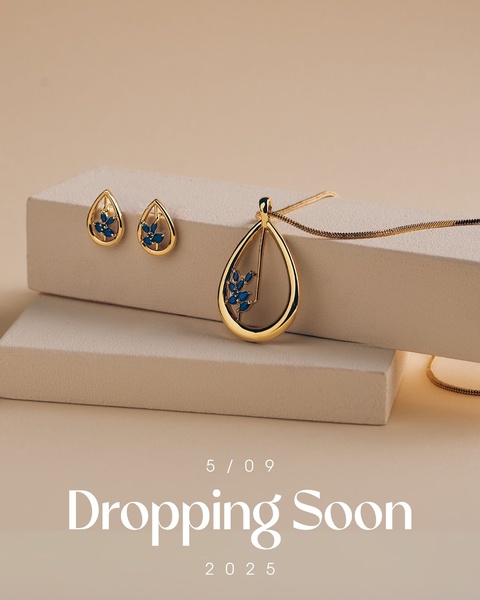 A high-end teaser image showcasing a gold teardrop pendant and matching stud earrings on a neutral platform, announcing a future product drop date.