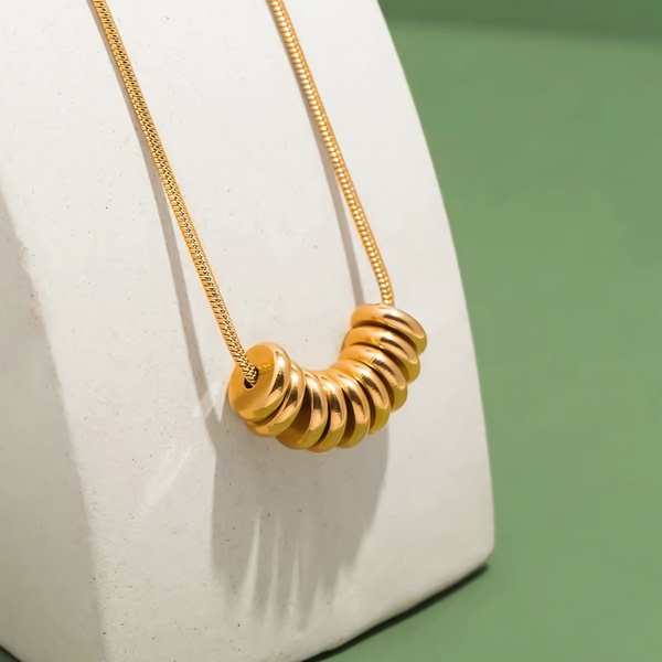 A high-end studio photograph featuring a gold necklace with multiple ring pendants, displayed on a clean white plinth against a sage green background.