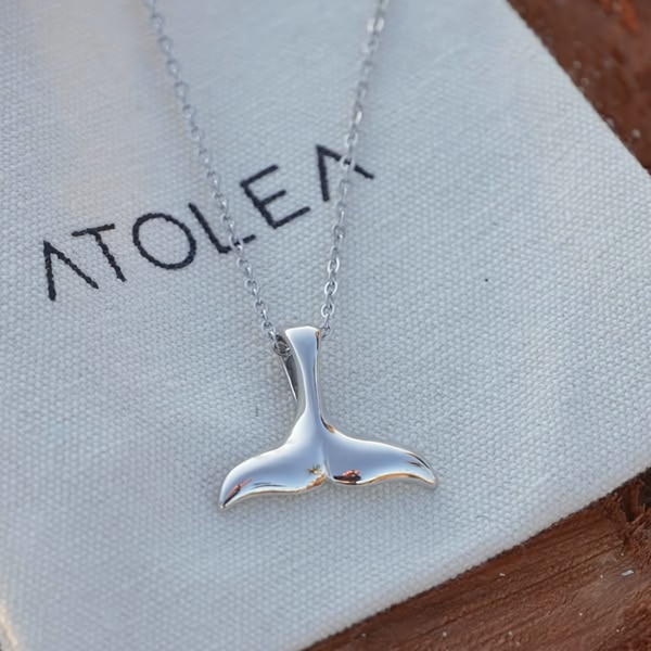 A close-up photograph of a silver whale tail necklace resting on a branded dust bag.