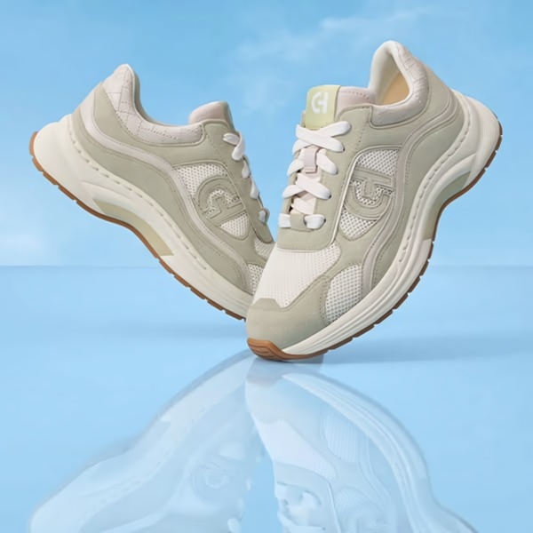 A high-end product hero shot featuring a pair of running shoes suspended over a reflective surface against a soft sky-blue backdrop.