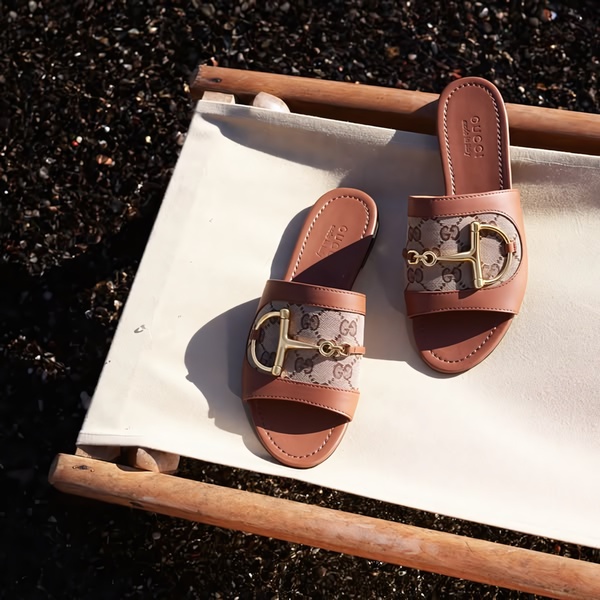 A high-end product hero shot displaying a pair of designer slide sandals resting on a clean canvas chair against a textured dark beach background.