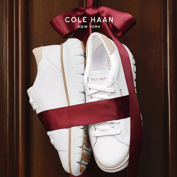 A pair of clean white sneakers tied together with a deep red satin ribbon hanging against a classic wood door background.