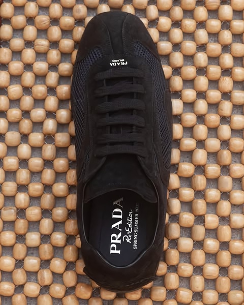 A top-down, centered view of a black suede Prada sneaker placed on a textured wooden bead mat.