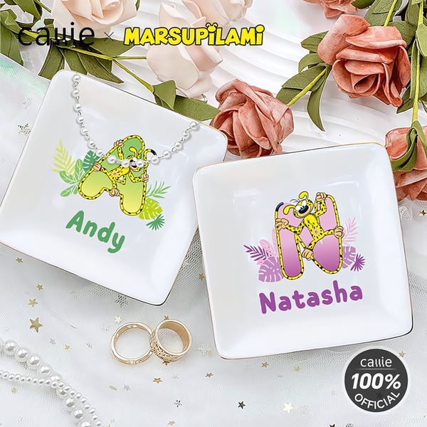 A high-angle product shot of two personalized jewelry dishes featuring character designs, surrounded by soft-focus flowers and delicate rings.