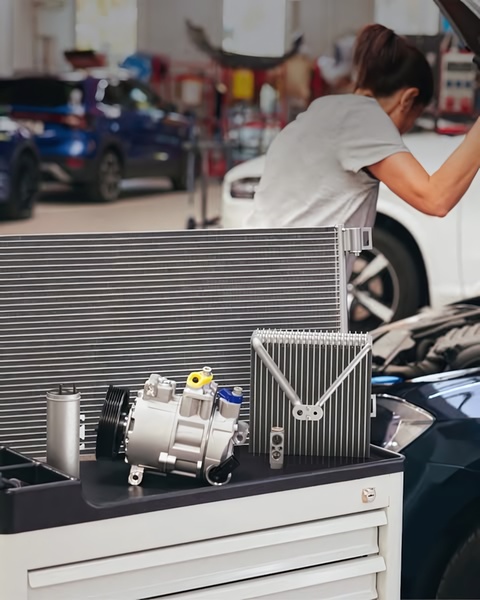 A high-quality studio-style product shot featuring a car AC compressor and radiator placed on a mechanic trolley, with a technician working in the blurred background.