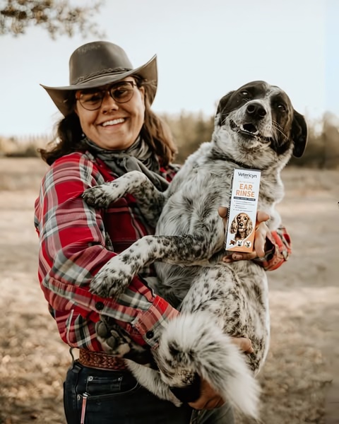 A pet owner wearing a cowboy hat poses in a natural, outdoor setting while holding a dog and a bottle of ear rinse.
